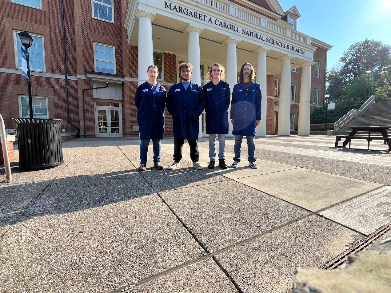 Four Berea College physics students stand shoulder to shoulder in front of the Margaret A. Cargill Science building wearing blue lab coats, facing the camera.