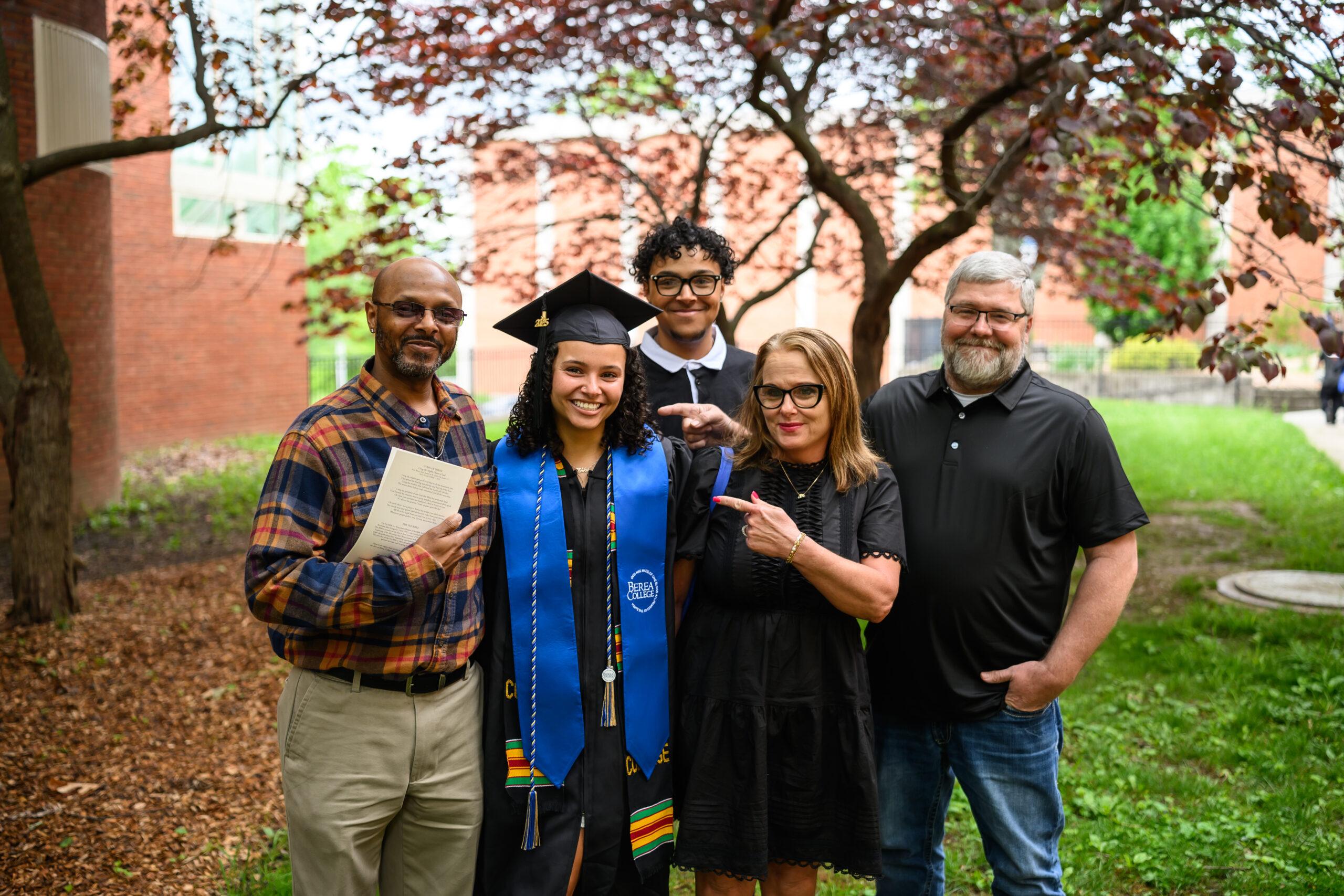 A photo of a student at their Berea graduation, in a traditional black gown and graduation cap, as they pose for a photo with their family who is pointing to the graduate and smiling proudly.